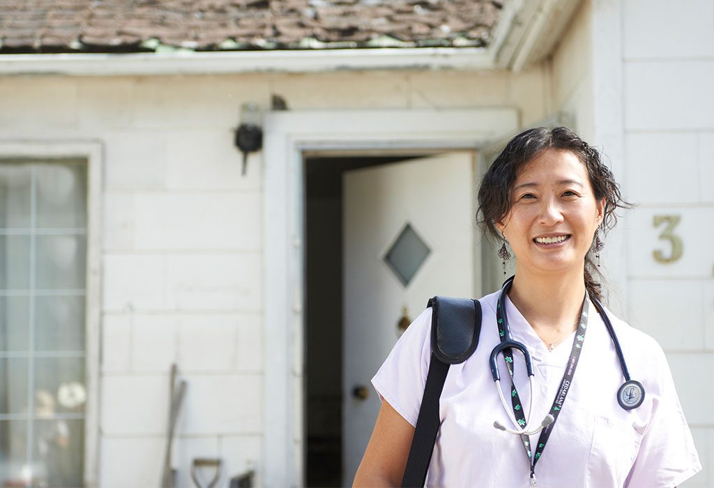 A medical professional wearing scrubs and a stethoscope poses and smiles in front of an open door.