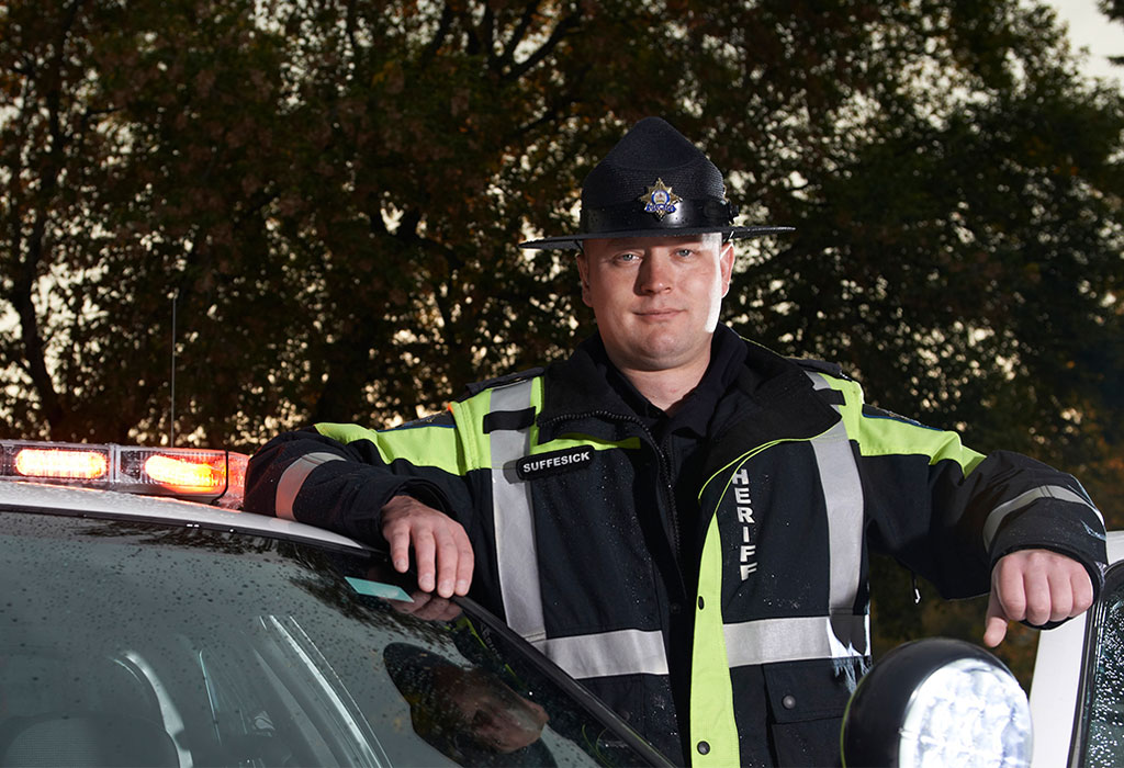 An AUPE Local 003 sheriff leans against a police car.