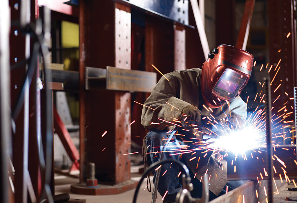 A welder, wearing a welding mask and personal protective gear, leans over their equipment with welding sparks flying.