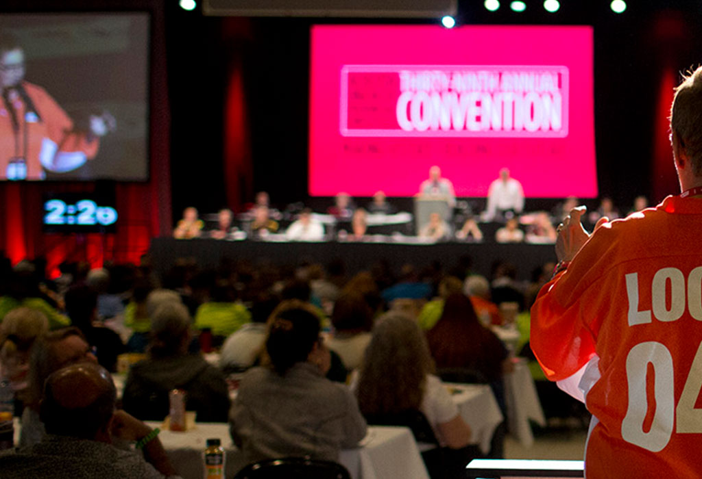 A delegate to Convention stands at the microphone stand and speaks to the other delegates on the Convention floor; the stage and screen as well as the rest of the Delegates are shown blurred in the background.