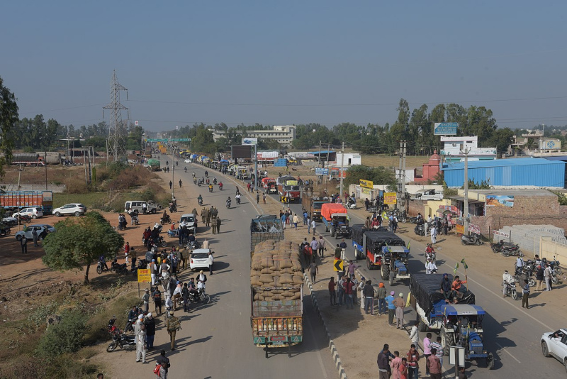A caravan of farmers drives down a road in India. Many of the vehicles have people filling the beds of the trucks, and farmers' flags hanging off the sides.