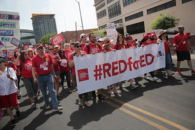 A large crowd of teachers, all wearing red, march behind a banner that reads "Red for Ed."