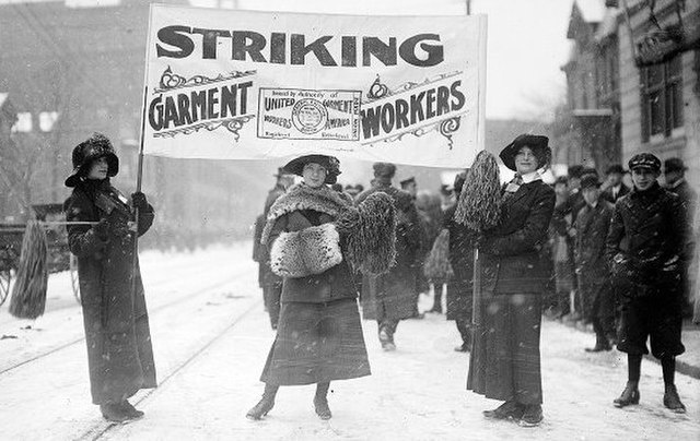 A black and white image of three women holding a banner which reads "striking garment workers" in front of a crowd of striking workers on a snowy day.