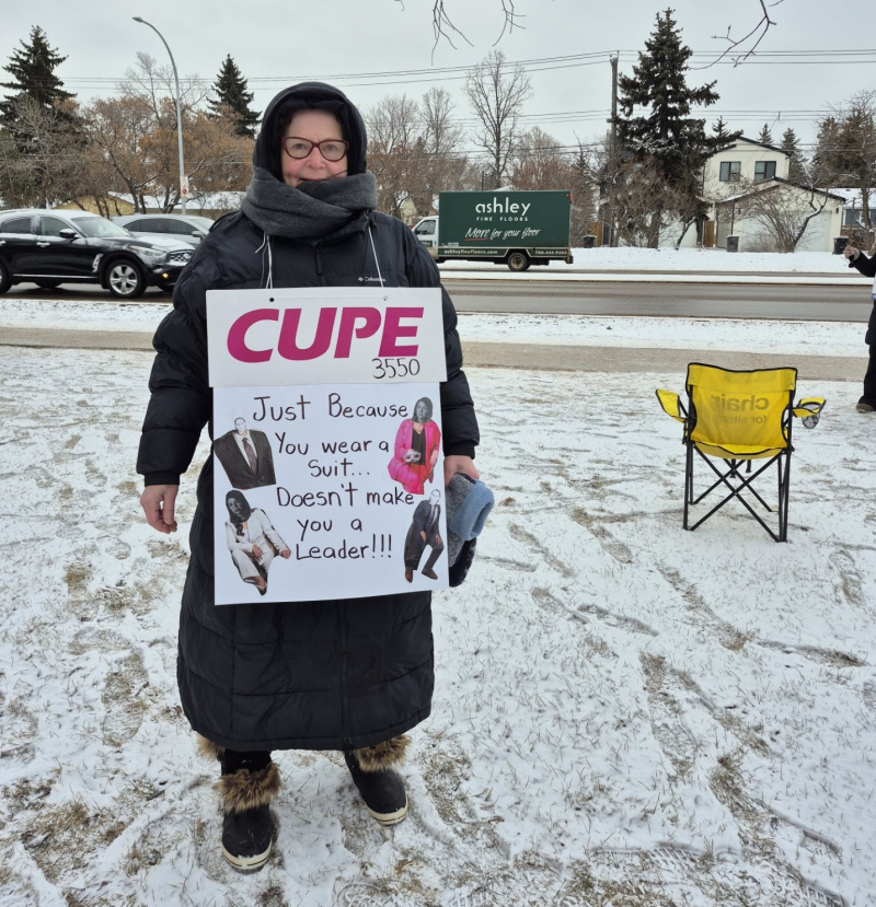 Pearl Biggar stands in a snowy field dressed in winter layers, a big scarf around her neck. Her sign reads, "CUPE 3550: Just because you wear a suit... Doesn't make you a leader!!!"