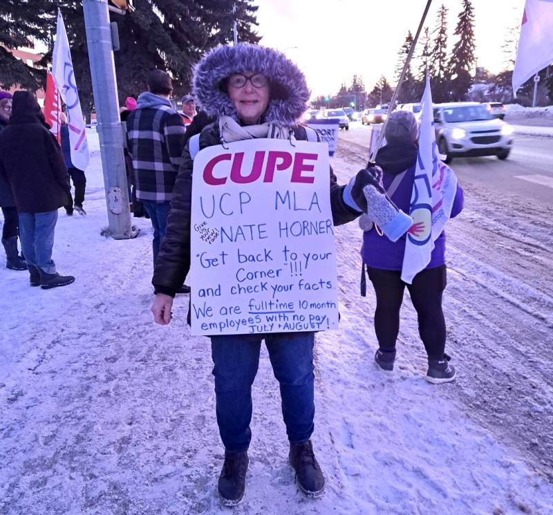 Pearl Biggar stands on another picket line, dressed in a winter jacket with the hood up. Her CUPE sign reads, "UCP MLA Nate Horner: Get back to your corner!!! and check your facts... We are full-time 10 month employees with no pay July + August"